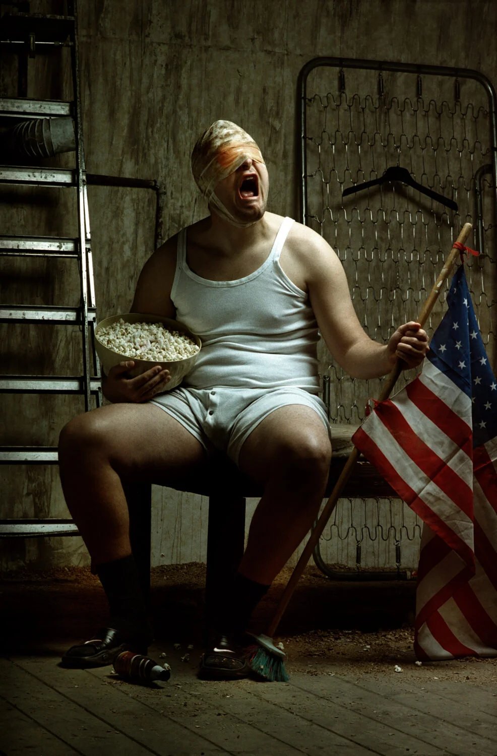 A man with a bandaged head sitting on a bench in a basement, holding a bowl of popcorn in one hand and an American flag in the other. The man appears to be shouting or crying, and there are shelves and a metal wall in the background.