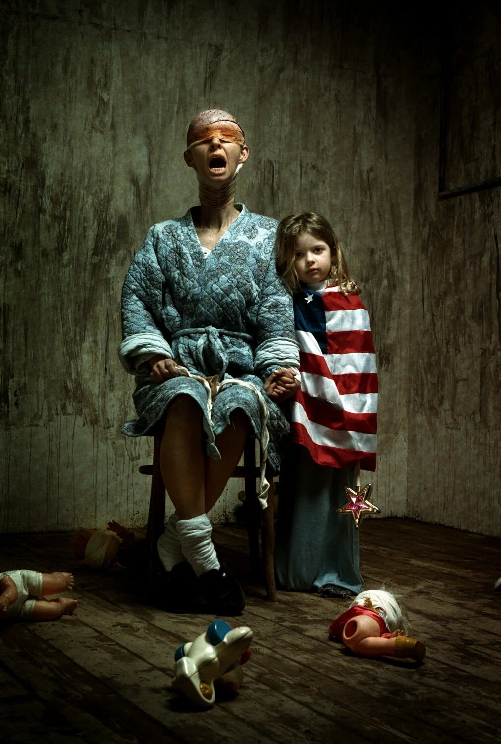 A woman with a blindfold sitting on a wooden chair, holding hands with a young girl who is standing beside her wearing an American flag cape. The setting is a dimly lit, worn room with toys scattered on the floor.