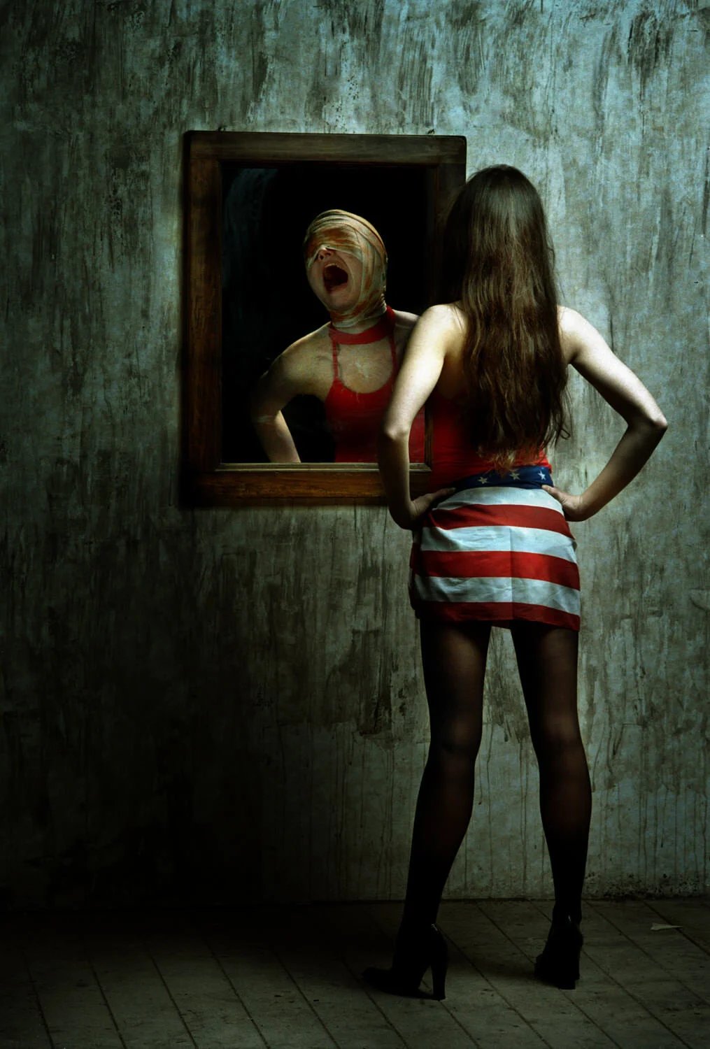 Young woman with long brown hair wearing a red top, American flag skirt, black stockings, and high heels standing with hands on hips looking into a mirror. The mirror reflects a woman with a blindfold over her eyes and a distressed expression, wearin