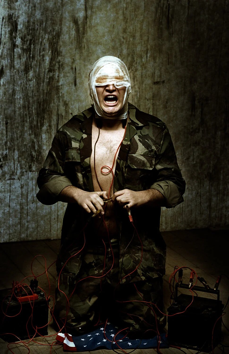 A man wearing camouflage pants and jacket, with his face wrapped in bandages, is yelling with his eyes blindfolded. He is standing shirtless on an American flag on the floor, surrounded by wires and batteries, in front of a textured wall.