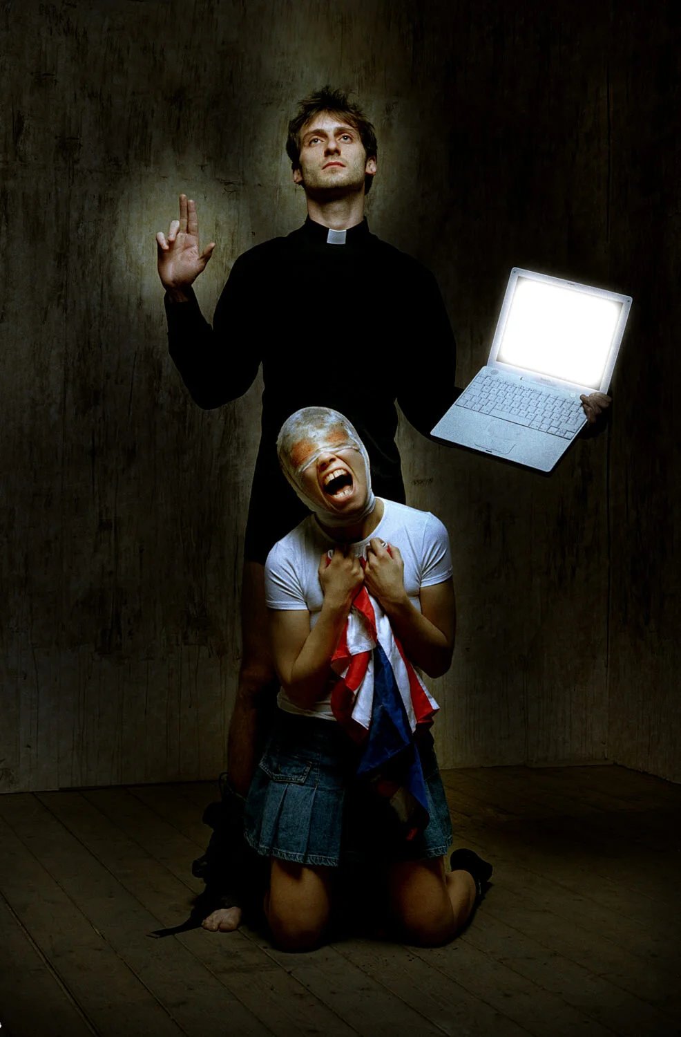 A man dressed as a priest holding a laptop with a bright screen, standing behind a woman kneeling on the floor with a distressed expression, holding a shirt with the American flag pattern.