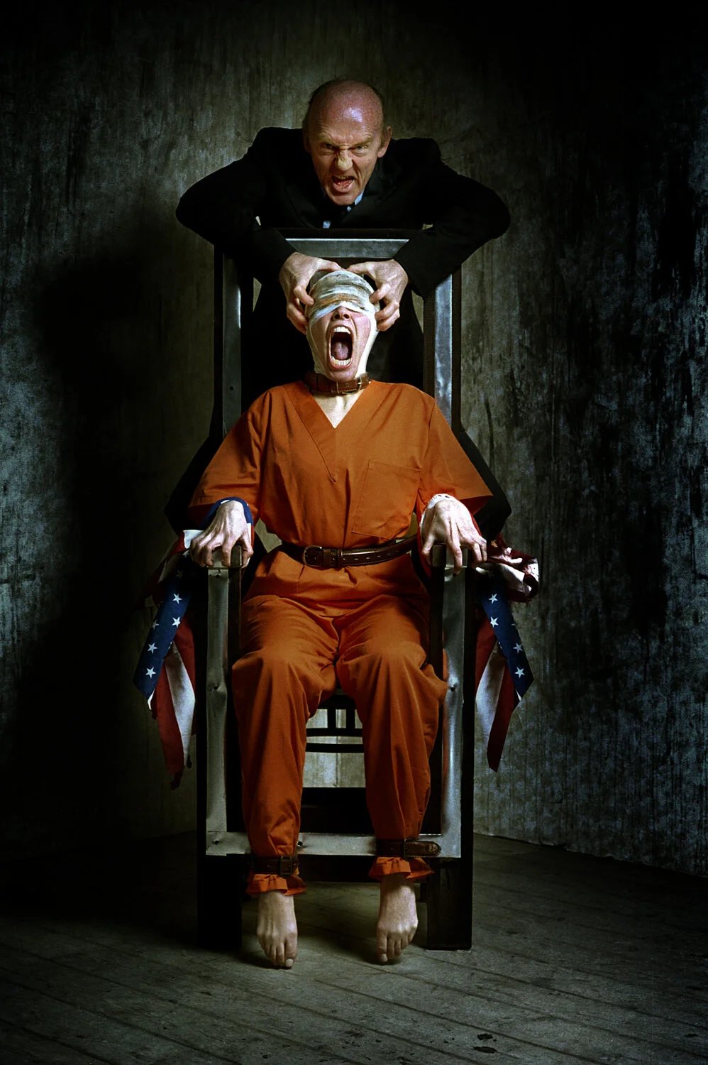 A person in an orange prison jumpsuit is tied to a chair with US flags on the sides, screaming with an open mouth while an older man stands behind them, pulling a blindfold off their eyes. The scene is dark and intense.
