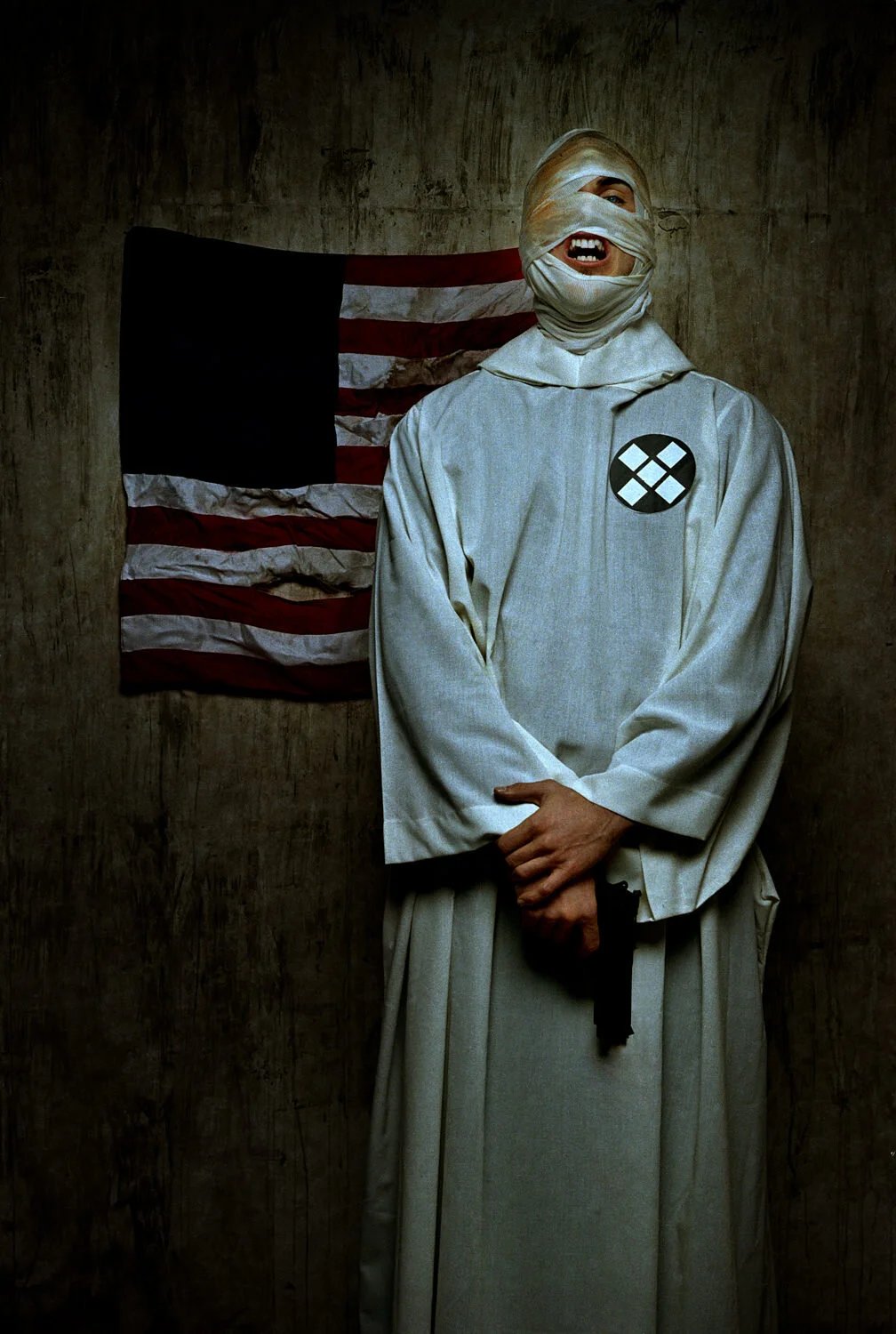A person dressed as the character the Nun from 'The Conjuring' series, with bandages wrapped around their face, standing in front of an American flag with a worn appearance, on a textured wall.