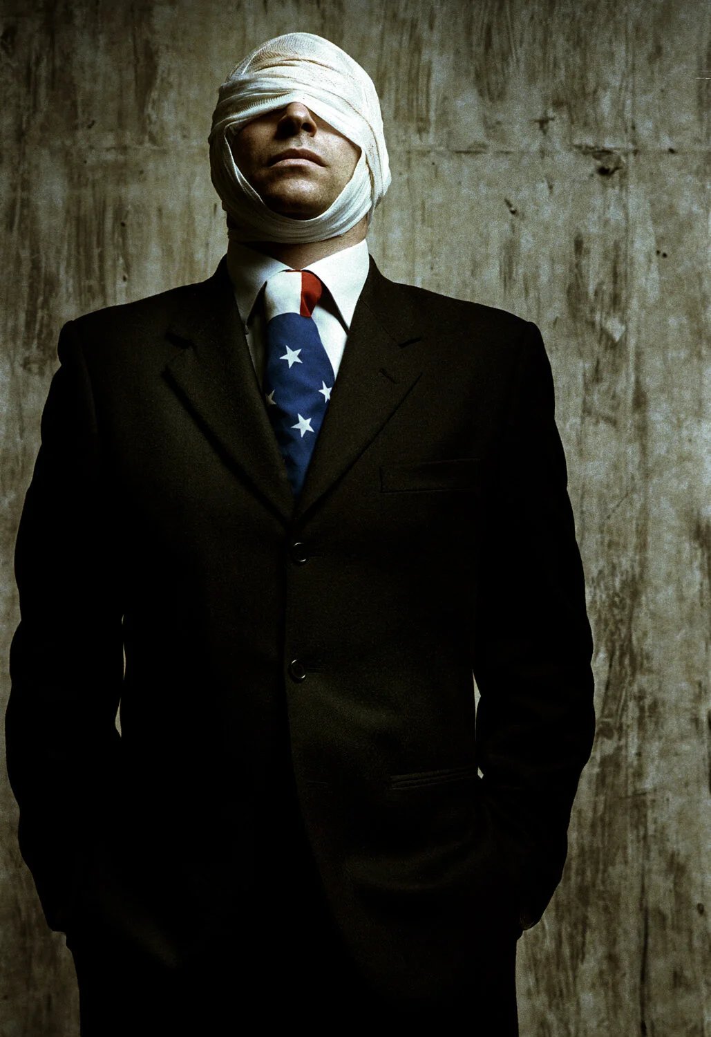 A man in a black suit and tie with an American flag pattern, his face and eyes covered with cloth, standing against a textured wall.