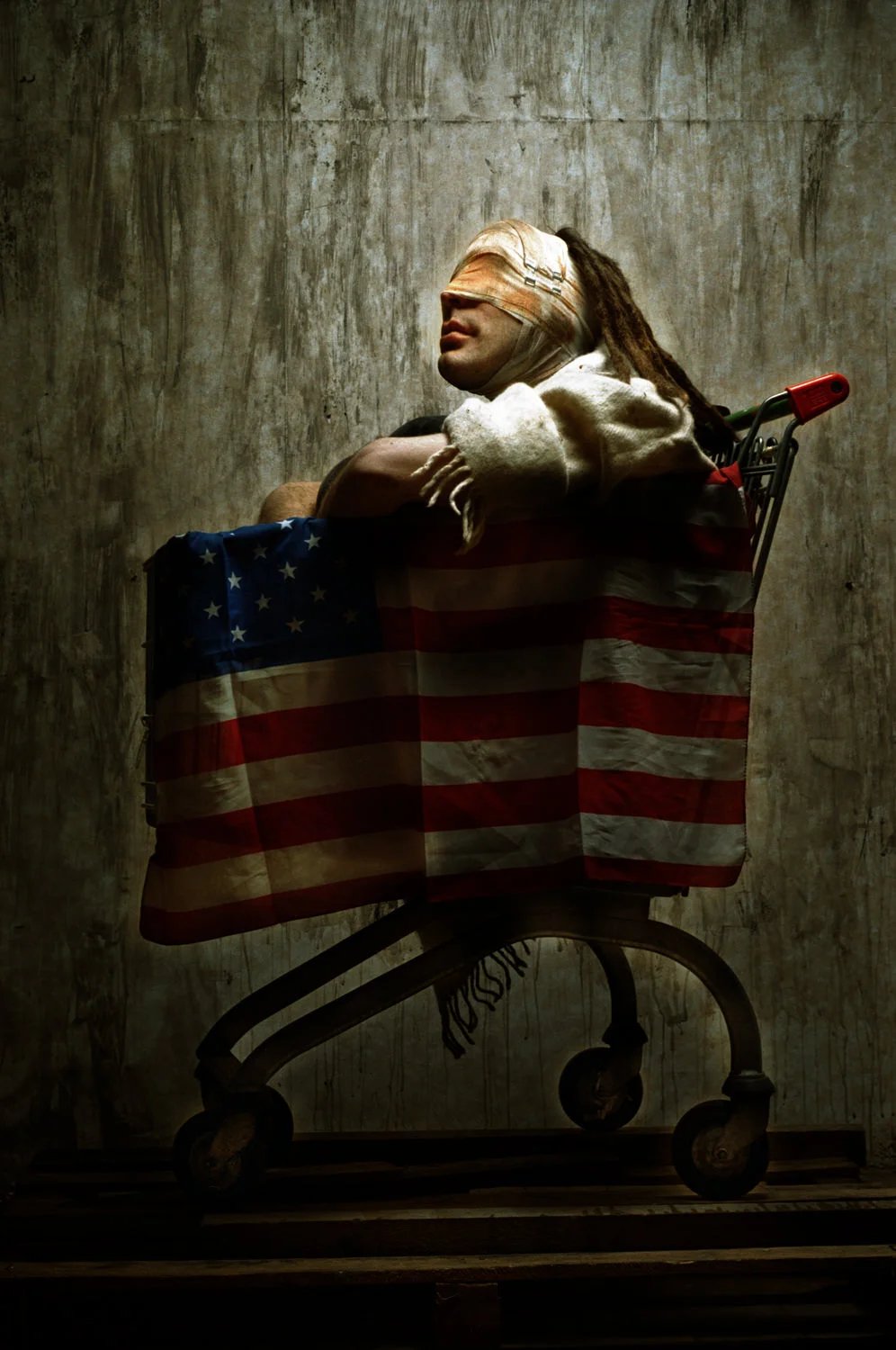A person with a bandaged head and dreadlocks sitting in a shopping cart draped with an American flag, in front of a textured, concrete wall.